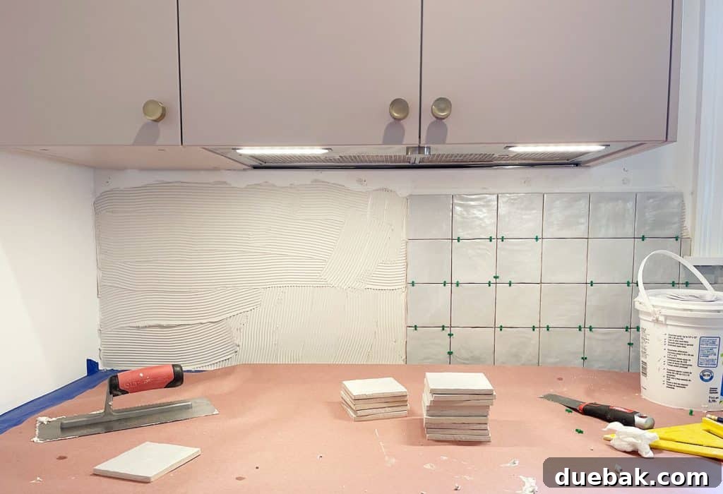 Person installing zellige-style tile backsplash under mauve cabinets, demonstrating the tiling process.