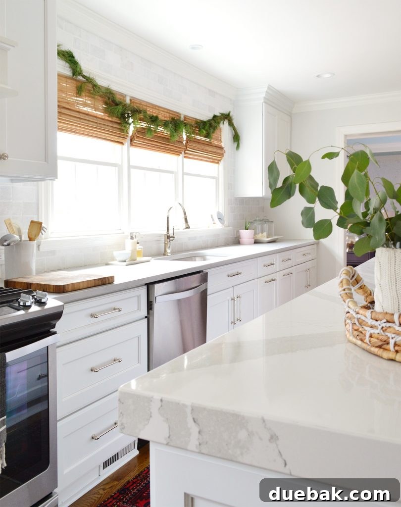 A pristine white kitchen featuring a festive fir garland draped elegantly over the sink windows, adding natural holiday charm to the space.