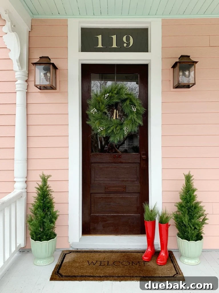 A charming pink house with a wooden door, festive with a faux Christmas wreath and symmetrical faux evergreen trees on both sides, creating a welcoming and elegant holiday facade.