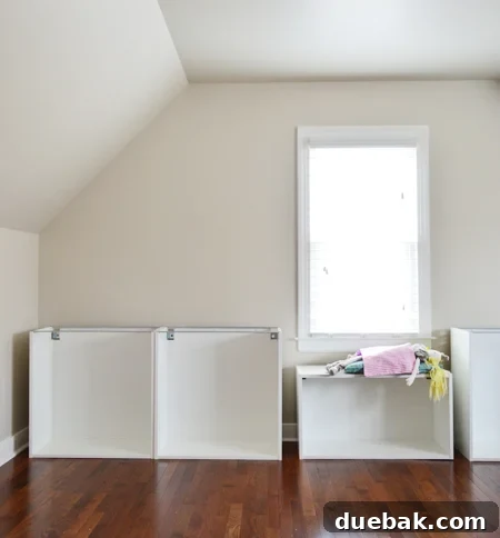 Crafting Custom Built-In Cabinetry 5 Bonus room with sloped ceiling, showing partially assembled Ikea cabinet boxes arranged in place, with a child's doll sitting on one, demonstrating the window seat concept