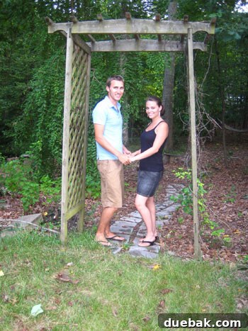 A couple playfully posing under a rustic wedding arch now moved to the entrance of their backyard woods, recreating a sweet moment from their wedding day for their anniversary.