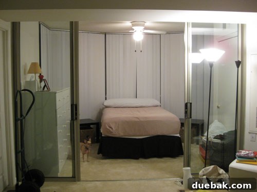 Before photo of a sunroom with a bed, showing expansive glass walls and vertical blinds, looking somewhat uninviting.