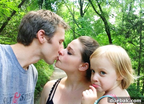 Candid family photo attempt with parents kissing and child making a funny face, showing the playful reality of family photos.