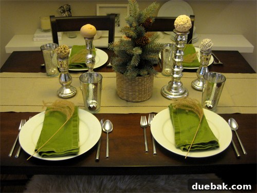 Elegant Thanksgiving table setting combining metallic silver cups and candlesticks with woven orbs, a faux-spruce tree nestled in a textured basket, refreshing green napkins, and delicate dried grass cuttings for an organic touch.