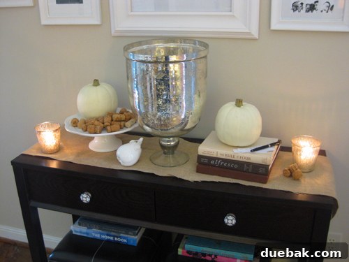 Close-up of a festive Thanksgiving console display, highlighting white pumpkins, shimmering votive candles, and a collection of natural wine corks artfully arranged on a stand.