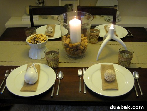 Modern and airy Thanksgiving table setting featuring a cork centerpiece, an elegant antler candle, an inverted scalloped bowl with caramel corn, woven cups, burlap napkins, ceramic pears, and decorative woven orbs.