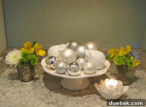 Frugal Florals for a Fresh Start 3 A close-up view of the New Year's themed floral arrangement, artfully combined with festive ornaments and ceramic fruit on a decorative cake stand, enhancing the home's winter ambiance.