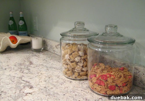 Countertop with two clear cereal jars, one filled with granola and the other with cheerios, next to a fresh bowl of cereal.