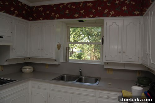 Transform Your Kitchen Without Breaking the Bank 2 Dated kitchen with yellow walls, dark wood cabinets, and a closed-off feeling before renovation. A cluttered counter space and older appliances are visible.