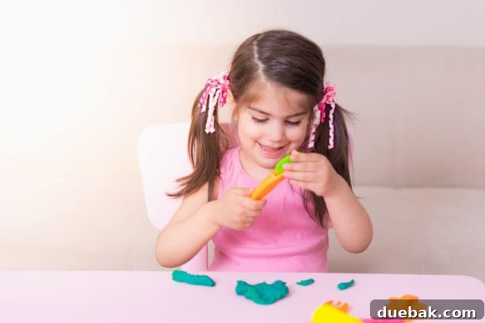 A cute young girl happily playing with colorful playdough on a mat near a table.