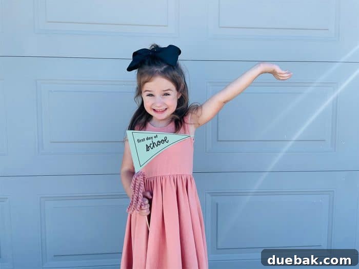 A cheerful girl in a pink dress proudly holding a free printable first day of school sign. The sign is a stylish triangle-shaped pennant banner, attached to a stick with decorative ribbons and bows, perfect for a photo op.