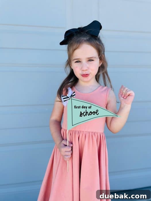 A young girl in a pink dress, smiling as she holds a free printable first day of school sign. The sign is a trendy triangle pennant banner, secured to a stick and embellished with delicate ribbons and bows, embodying a perfect photo prop.