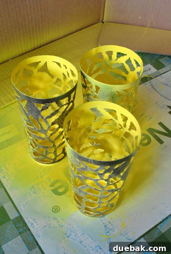A person's hands holding a metal cup as the interior is being spray painted yellow, with some overspray visible on the outside. A protective mat is underneath.