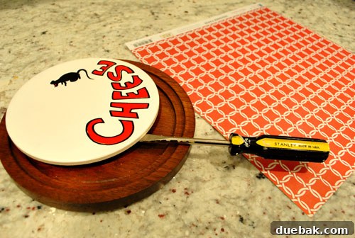 Close-up of a flat-head screwdriver prying the ceramic insert from the wooden base of a cheese plate.