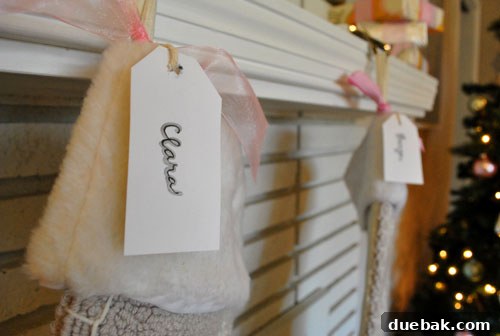 Personalized card stock tags, tied with ribbon, hanging from Christmas stockings on a fireplace.