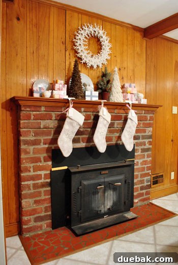 Decking Our New Home for Christmas 4 The kitchen side of a fireplace, featuring dark paneling, brick, and red tile, adorned with various holiday decorations for a festive look.