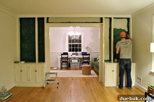 Transform Built In Bookcases With A Painted Back 2 Man wearing a 'Talk Nerdy To Me' shirt, standing in front of partially painted built-in bookshelves during a home renovation project. The shelves are in a transitional state, showcasing the DIY process.