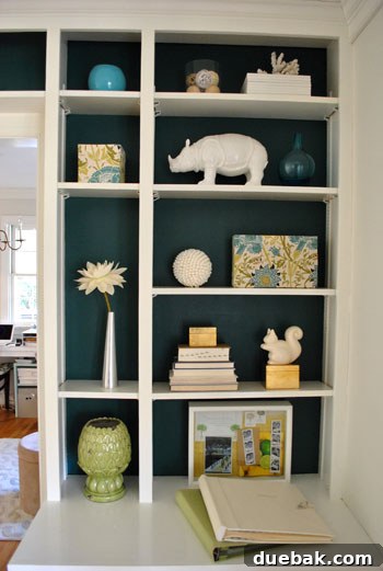 Transform Built In Bookcases With A Painted Back 5 Vertically oriented image showcasing a styled built-in shelf. Various white ceramic objects and decorative items pop elegantly against the newly painted deep blue-green backdrop, emphasizing contrast.