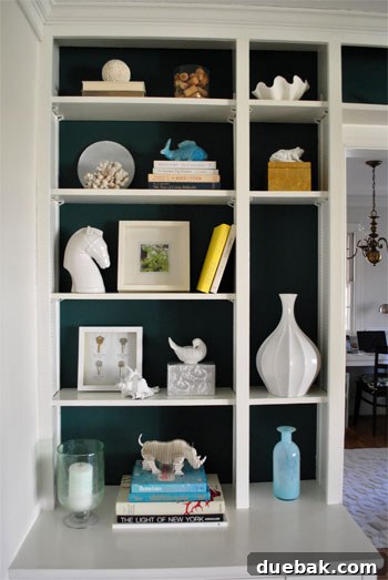 Transform Built In Bookcases With A Painted Back 6 Another vertical shot of a beautifully styled bookshelf. A collection of white decorative objects, including ceramics and unique finds, contrasts sharply and elegantly with the dark blue-green paint.