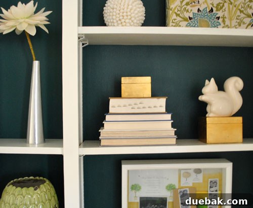 Transform Built In Bookcases With A Painted Back 7 Wide shot of the finished built-in bookshelves, elegantly styled with books, various decorative items, and white ceramics. The rich blue-green color of the shelves creates a sophisticated and cohesive living room focal point.