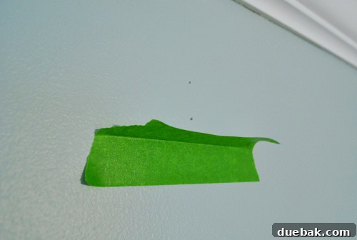 Dust-Free Drilling with Painter's Tape 2 Close-up of painter's tape folded to create a dust collection shelf below a drill mark on a wall, preventing dust from falling to the floor.