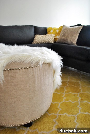 Large round upholstered ottoman with nail head trim atop a yellow Pottery Barn rug, adding a soft contrast to the sectional.