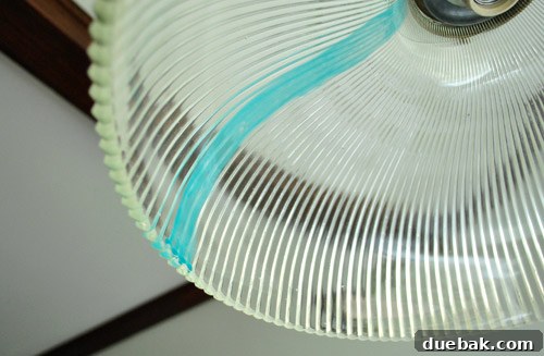 A hand using a sponge brush to carefully paint the inside of a glass light shade with diluted turquoise acrylic paint, creating vertical stripes.