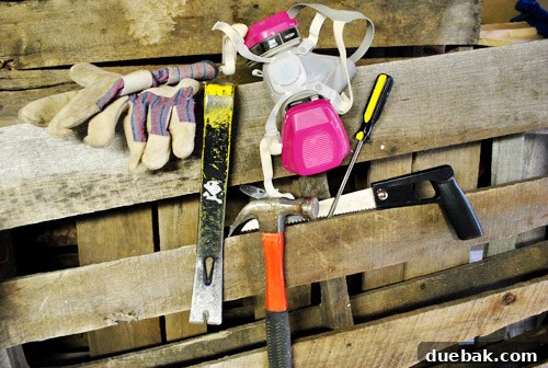 DIY tools laid out for pallet deconstruction: crowbar, hammer, saw, gloves, mask