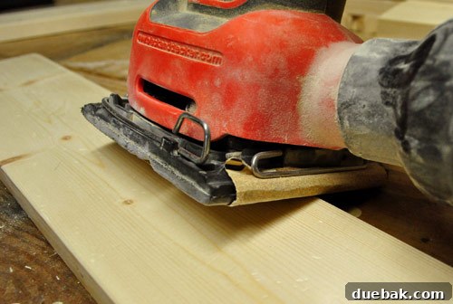 Using a black & decker palm sander to gently round and smooth the sharp edges of a new wood board, an initial step in creating an aged and distressed look.
