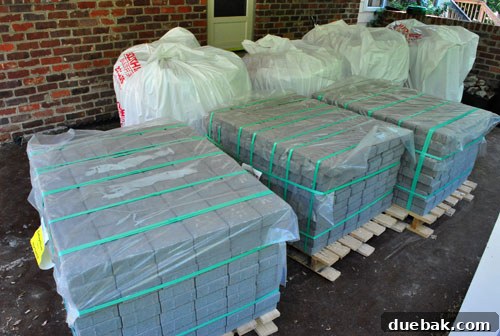 Patio construction materials neatly stacked in the carport: bags of gravel and sand, and pallets of cobblestone pavers