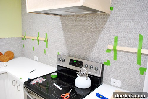 Scrap wood pieces taped to the tiled kitchen wall as temporary guides for bracket placement.