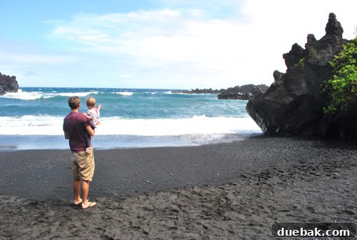 Clara, our toddler, plays on the unique black sand beach at Waianapanapa State Park, with dark volcanic rocks and crashing waves.