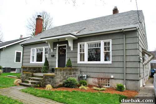 Wider shot of the Portland house exterior, nestled amidst existing greenery, hinting at spring/summer splendor