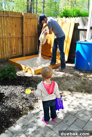 Clara patiently waiting with her bucket and shovel beside the partially filled sandbox