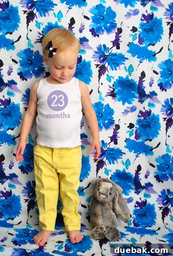Clara, 23 months old, posing in front of a neutral backdrop, smiling playfully for her monthly photo.