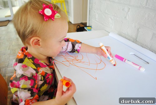 Clara, a toddler, is intensely focused on drawing with markers on a large sheet of paper spread on the floor.