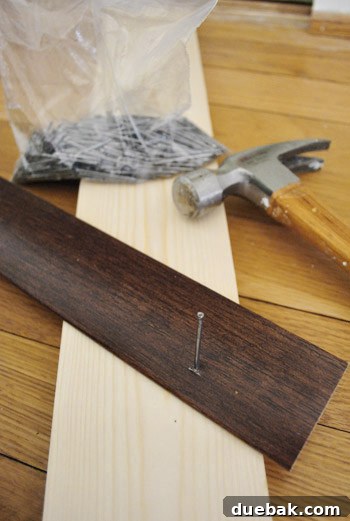 Close-up of a hand using a hammer to pre-set a nail into a wooden transition piece, supported by scrap wood.