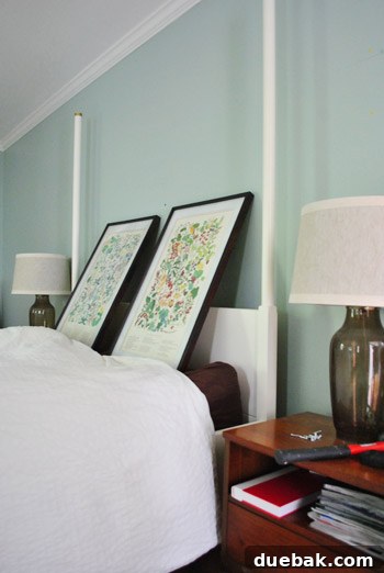 Two framed botanical prints propped up against a bedroom wall above a bed, in a trial placement before hanging.