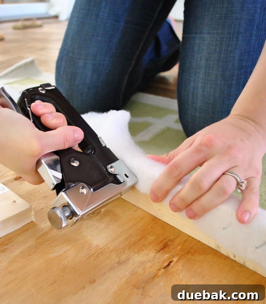 Using staple gun to attach batting to back of DIY upholstered fabric headboard, showing the stapling process