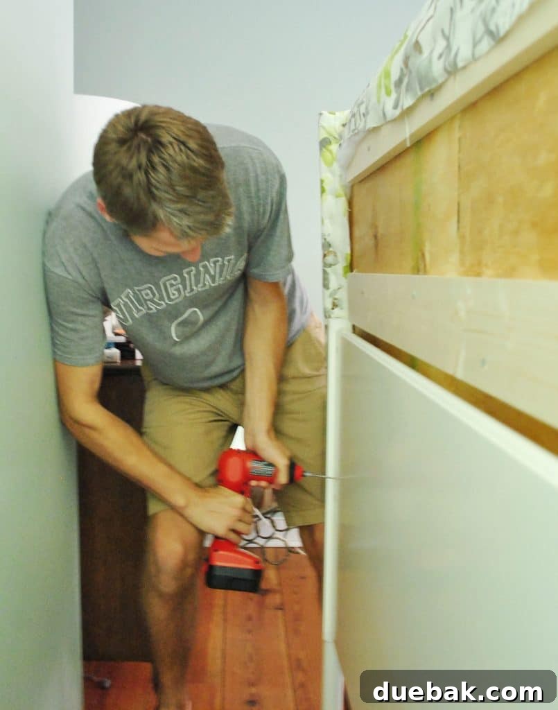 John screwing DIY upholstered headboard onto frame from backside of bed, showing the attachment process