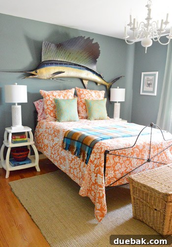 Guest room with dusty grey-blue walls, orange patterned bedding, a large thrift store sailfish, sculptural side tables, and textured lamps