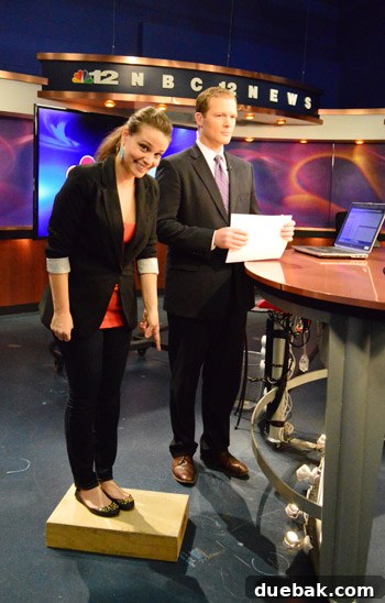 Sherry Petersik standing on a hidden box behind the news desk, smiling broadly next to John.