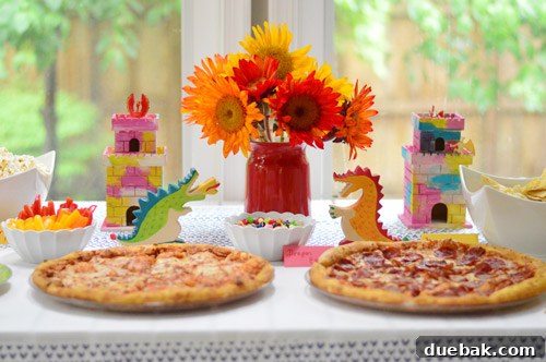 Dragon's Lair Birthday Bash 2 Dragon Themed Kids Party Food Display With Pizzas, popcorn, fruit, and snacks, surrounded by bright party decorations.