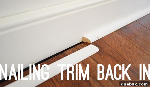 A nail gun being used to reinstall baseboards, with new quarter round visible on the newly installed hardwood floor.