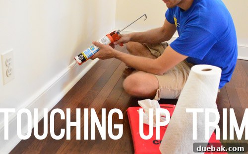 A hand touching up painted trim with caulk and paint, showing the final finishing touches after baseboard installation.