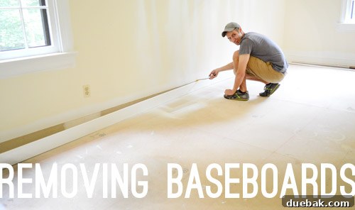 A crowbar being used to carefully remove baseboards from a wall, showing the utility knife score line for paint prevention.