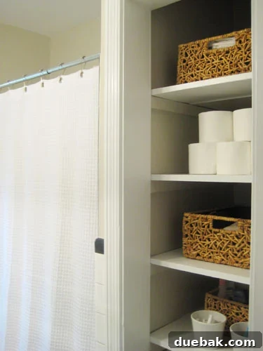 Neatly stacked toilet paper rolls forming a pyramid on an open shelf within a white bathroom linen closet, demonstrating organized visible storage