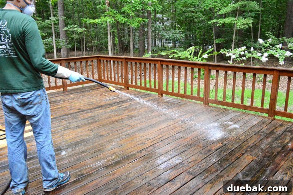 A person rinsing deck cleaner off a wood deck with a garden hose, showing clean water flowing