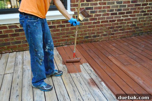 A person applying deck stain liberally with a roller on a wooden deck floor, working in manageable sections.
