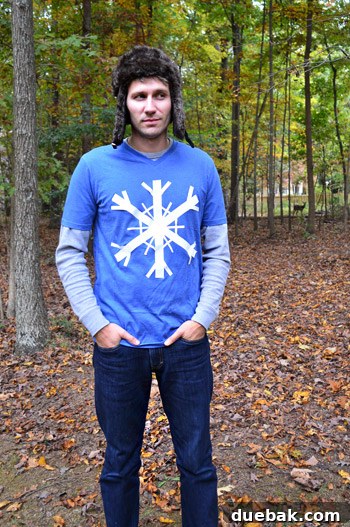John dressed in a blue t-shirt with a white duct tape snowflake, a wooly hat, standing in a field with a deer in the background.